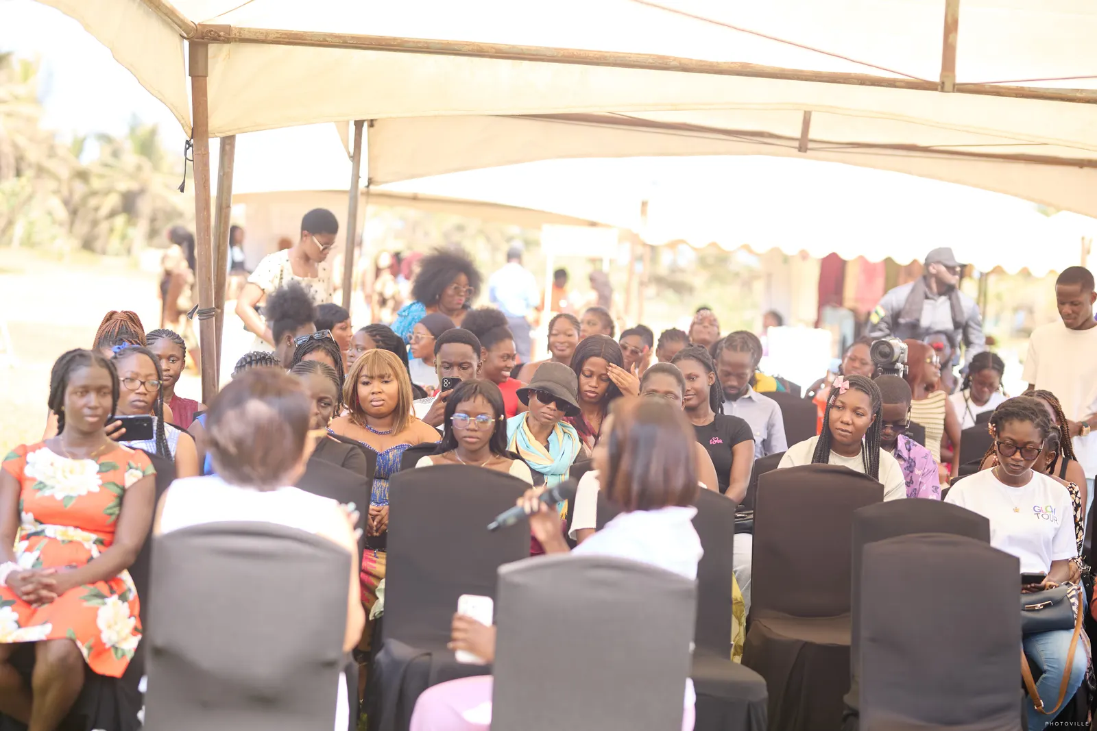 Large audience seated under white canopies at a daytime session