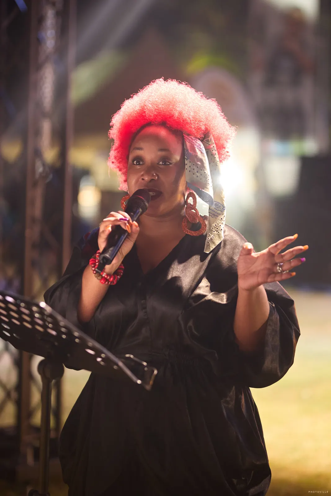 Performer with a red-tinted afro singing into a microphone at a music stand