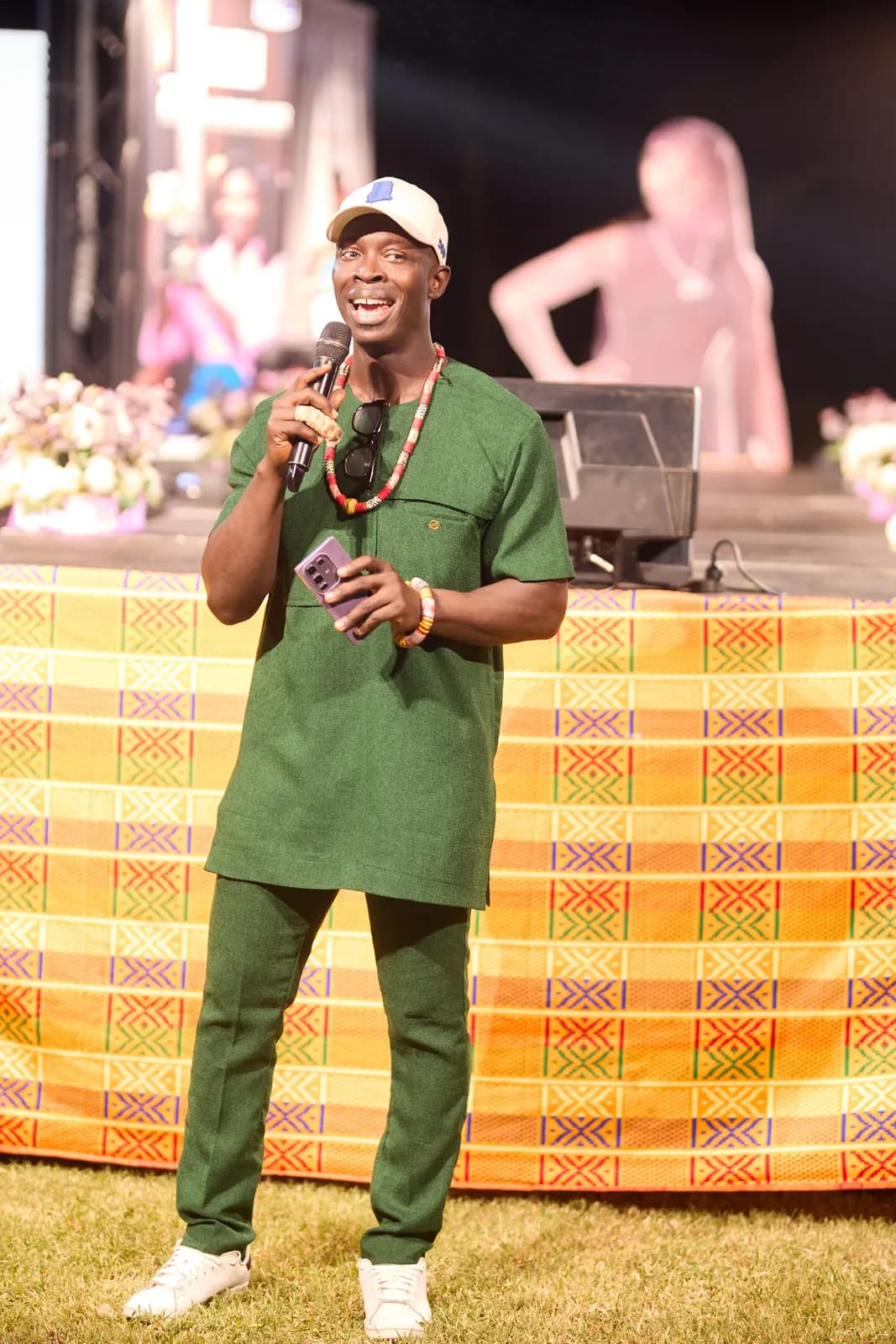 MC in a green outfit speaking on stage in front of a kente-draped table