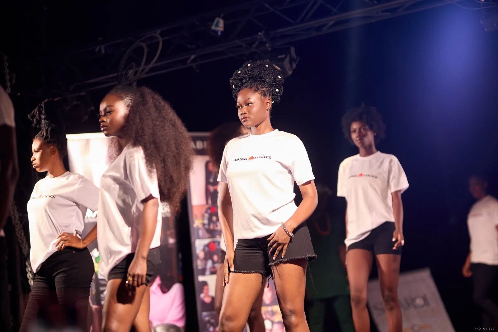 Models in white T-shirts with dramatic natural hairstyles posing on stage