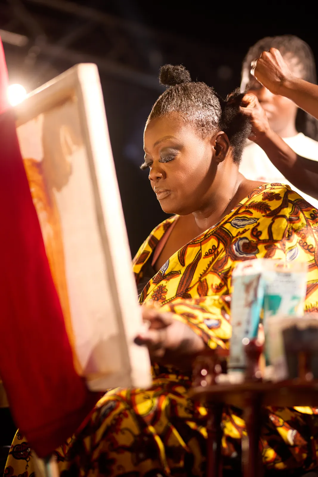 Artist in a yellow and black print outfit painting on stage while her hair is styled