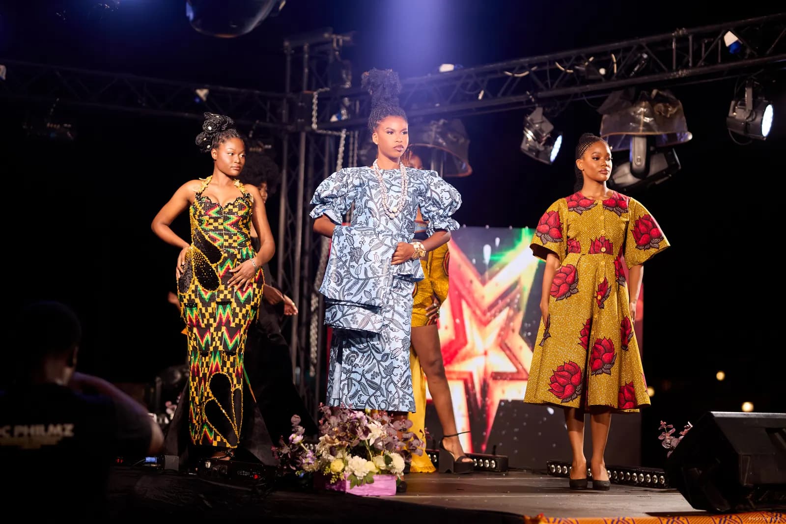 Three models in African print designs posing together on a lit stage